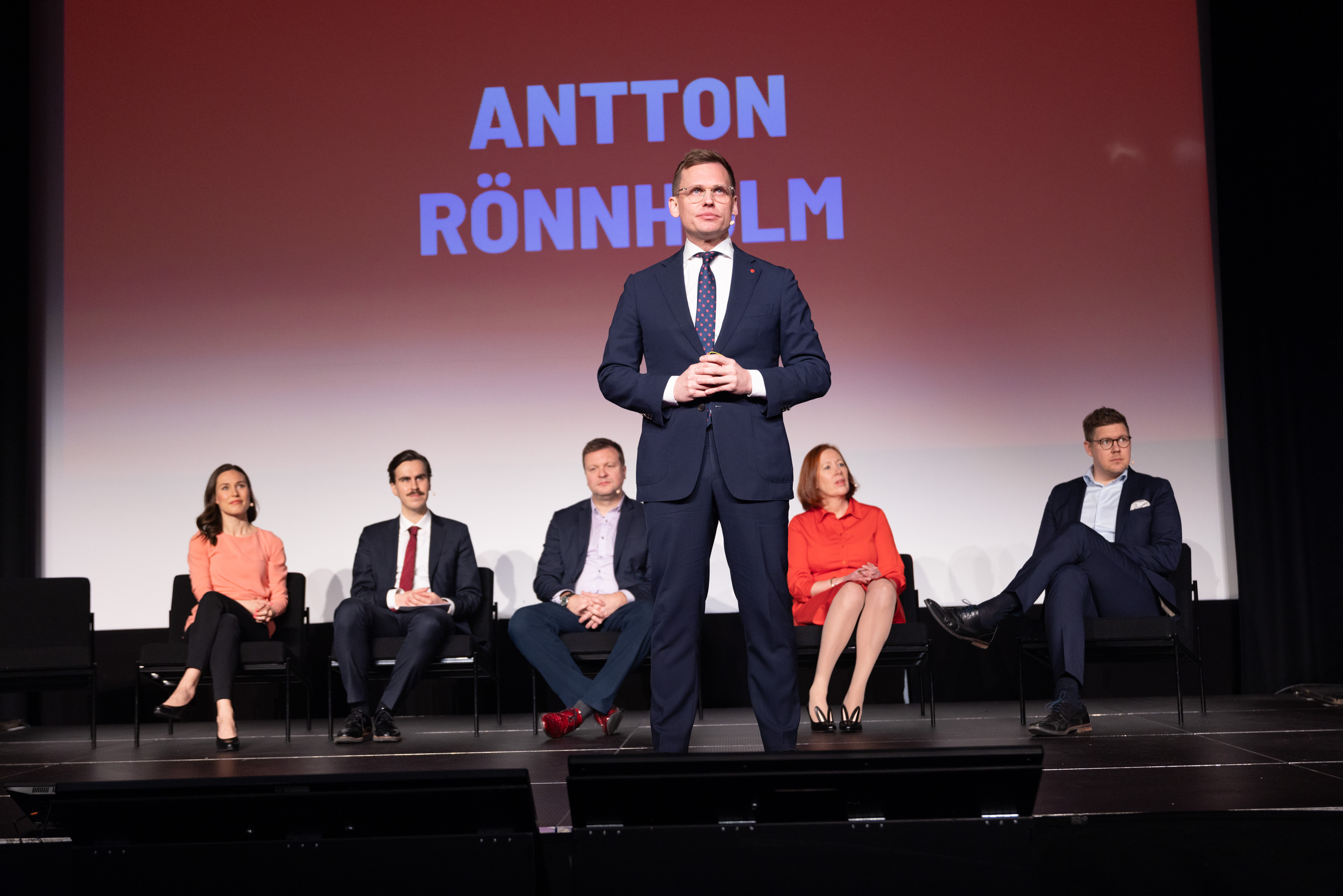 Antton Rönnholm puhumassa lavalla SDP:n tilaisuudessa, nimi taustanäytöllä